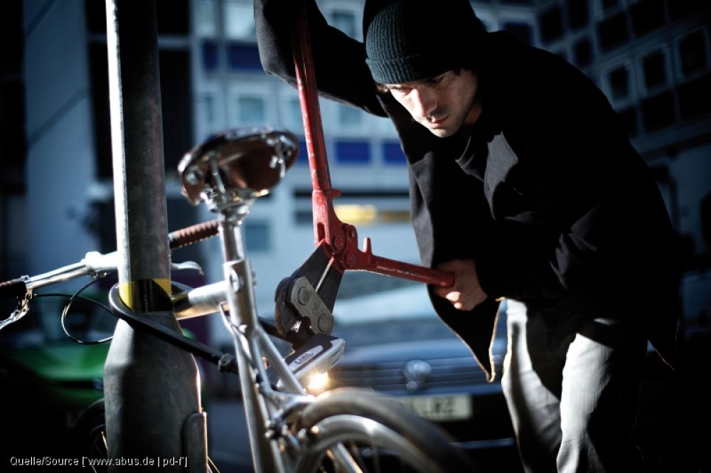 Ein Mann in dunkler Kleidung und dunkler Mütze versucht mit einem Seitenschneider ein massives Bügelschloss zu knacken. Das Rad ist an einem Metallrohr angeschlossen.